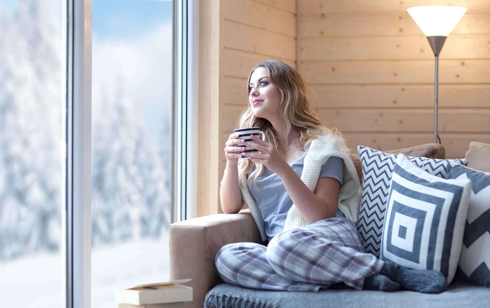 Young beautiful blonde woman with cup of coffee sitting home in living room by the window. Winter sn