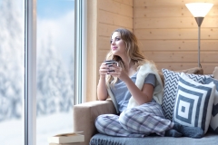 Young beautiful blonde woman with cup of coffee sitting home in living room by the window. Winter sn
